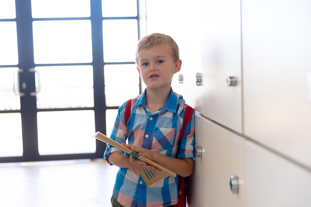 Portret van een schattige blanke basisschooljongen met een boek bij lockers op school