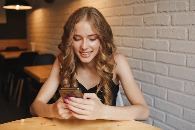 Portret van een schattige blonde vrouw die iets op haar smartphone leest terwijl ze in een moderne coffeeshop zit