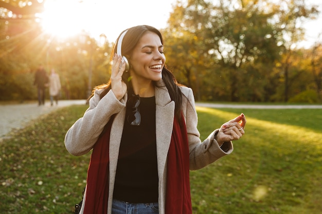 Portret van een schattige brunette vrouw met een jas die naar muziek luistert met een koptelefoon en zingt tijdens het wandelen in het herfstpark