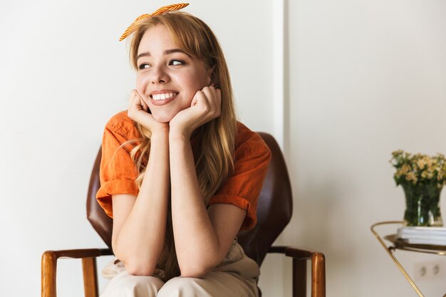 Portret van een schattige gelukkig optimistisch lachende jonge blonde vrouw thuis binnenshuis poseren zittend op een stoel.