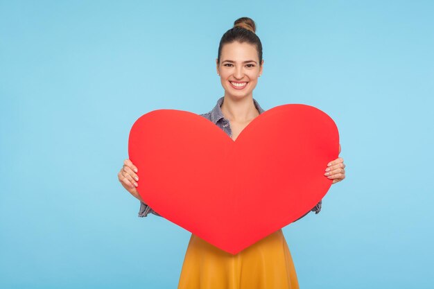 Portret van een schattige, gelukkige aantrekkelijke vrouw met een beminnelijke glimlach die een groot rood papieren hart vasthoudt, liefde genegenheid uitdrukt, romantische groet op Valentijnsdag. indoor studio-opname geïsoleerd op blauwe achtergrond