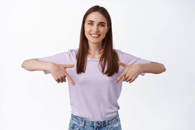 Portret van een schattige gelukkige vrouw die naar beneden wijst, witte tanden glimlacht en onderstaande advertentie toont, wijs naar het richtingslogo of banner, staande geïsoleerd tegen een witte achtergrond