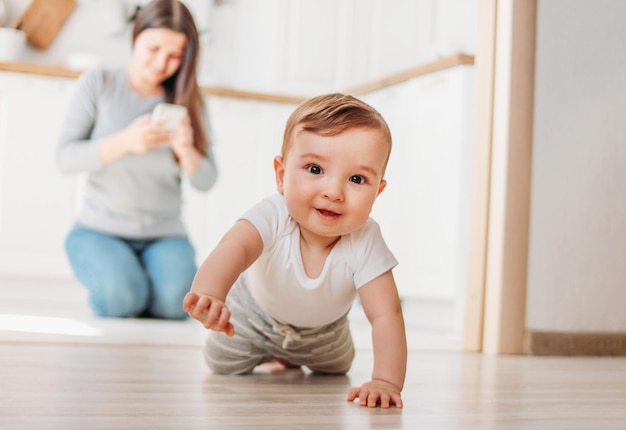 Portret van een schattige glimlachende baby jongen die tegen zijn moeder kruipt met zijn telefoon op de parketvloer thuis