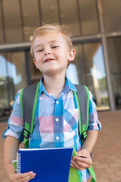 Foto portret van een schattige glimlachende blanke basisschooljongen met boeken die op de schoolcampus staan