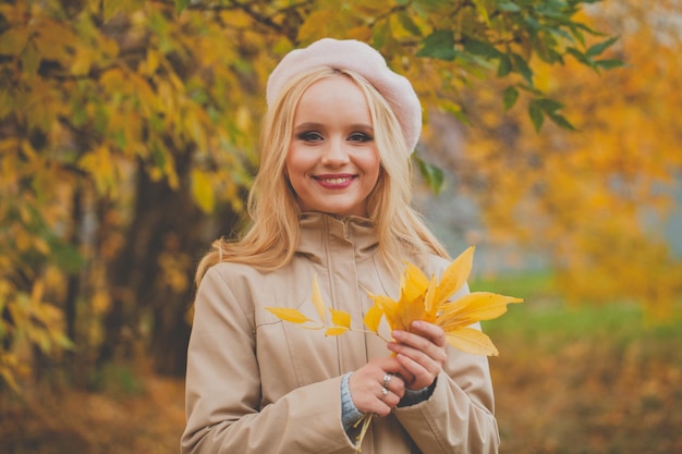 Portret van een schattige jonge gelukkige blonde vrouw die een Franse baret draagt met herfstbladeren buiten.