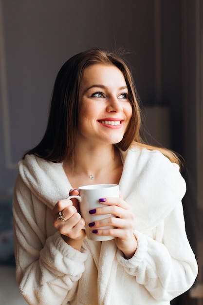 Portret van een schattige jonge vrouw die van hete thee geniet close-up. Vrouw houdt een kopje thee of koffie. Een gelukkig meisje droomt en kijkt naar het raam. Perfecte ochtend thuis.