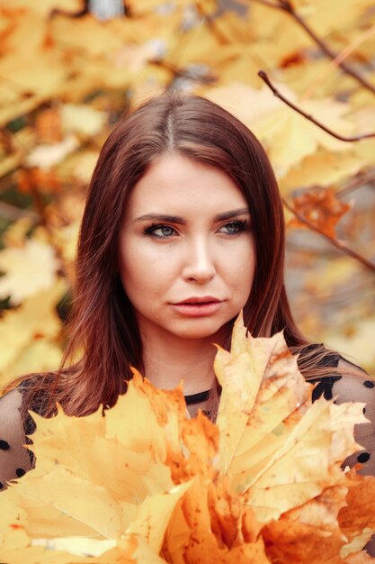 Portret van een schattige jonge vrouw met een Slavisch uiterlijk met bladeren in vrijetijdskleding in de herfst, staande tegen de achtergrond van een herfstpark. Vrij vrouwtje wandelen in Park in gouden herfst. Ruimte kopiëren