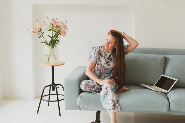 Portret van een schattige jonge vrouw met laptop die thuis werkt terwijl ze op een blauwe bank zit in een gezellige kamer