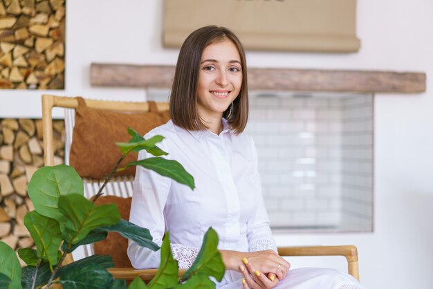 Portret van een schattige jonge vrouwelijke psycholoog in een gezellig licht kantoor zit in een fauteuil naast de binnenf...