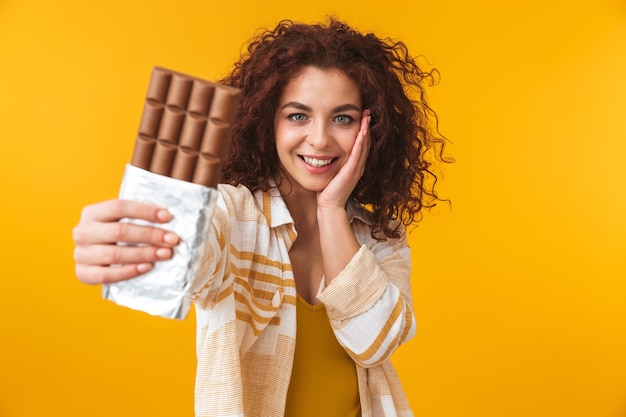 Portret van een schattige mooie jonge krullende vrouw die zich voordeed op een gele muur met chocolade.