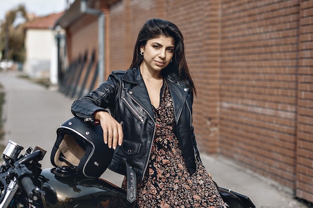 Portret van een schattige schattige brunette vrouw die zich voordeed op haar krachtige zwarte motorfiets, leunend tegen een zwarte helm in een jurk en leren jas. Aandrijfconcept