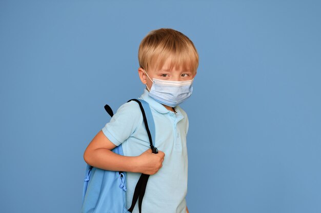 Portret van een schattige schooljongen jongen van 6 jaar oud met een lichte rugzak op een blauwe achtergrond. Terug naar school.