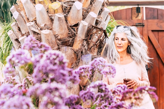 Portret van een schattige volwassen blanke vrouw met lang wit haar, trendy die een natuurlijke thee drinkt in een tuin buiten huis