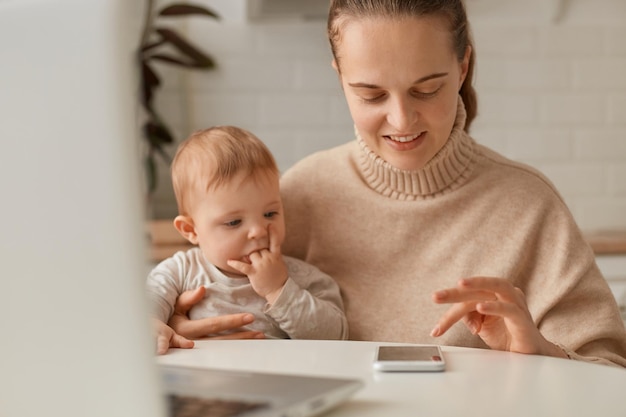 Portret van een schattige vrouw met donker haar met een beige trui die in de keuken zit en op een laptop werkt met een mobiele telefoon en een baby in handen houdt