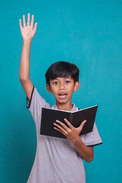 Portret van een schooljongen die zijn hand opsteekt en boeken vasthoudt die over groene achtergrond worden geïsoleerd