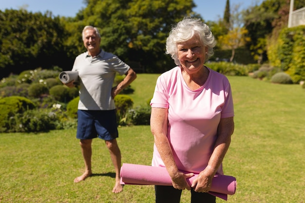 Portret van een senior blank paar dat yogamatten vasthoudt, naar de camera kijkt en glimlacht in de zonnige tuin. pensioenretraite en actief senior lifestyle-concept.
