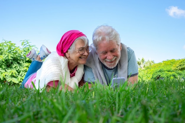 Portret van een senior glimlachend paar dat op het gras in een openbaar park ligt en samen naar een mobiele telefoon kijkt romantisch paar dat van vrije tijd en pensioen geniet