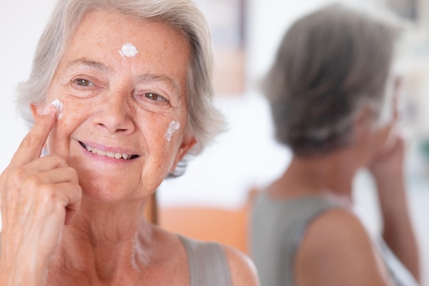 Portret van een senior mooie grijsharige vrouw brengt anti-verouderingscrème aan op een gerimpeld gezicht en zorgt voor het huidconcept