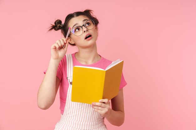 Portret van een serieus charmant meisje met een bril die een werkboek vasthoudt en geïsoleerd denkt over een roze muur