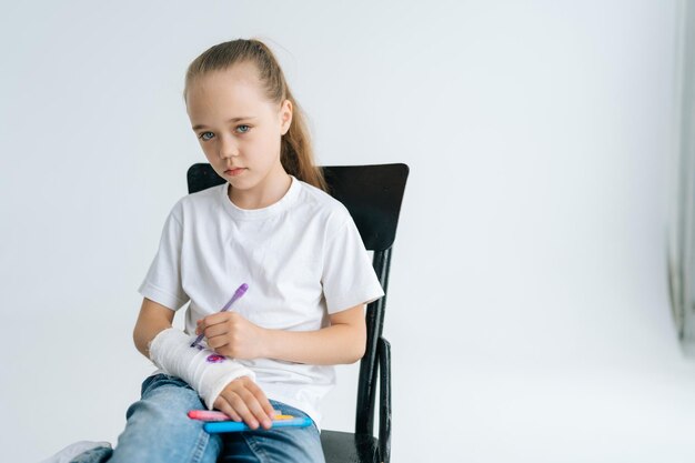 Portret van een serieus klein meisje dat een schattige afbeelding tekent met een kleurrijke stift op een gebroken arm, gewikkeld in wit gipsverband en kijkend naar de camera