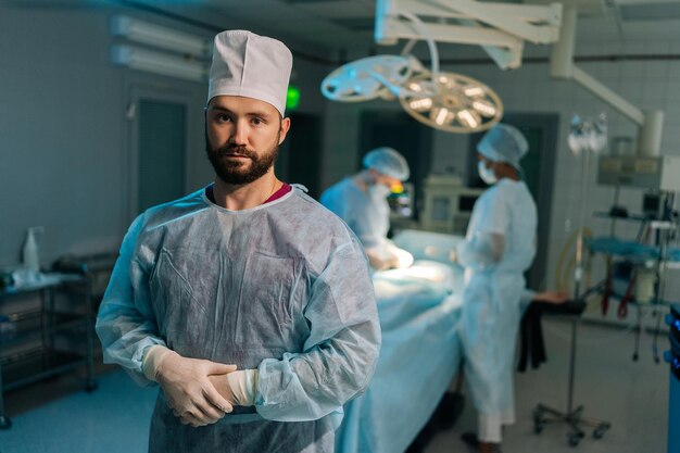 Portret van een serieuze baardige mannelijke arts in chirurgische uniformen die opstaat en naar de camera kijkt