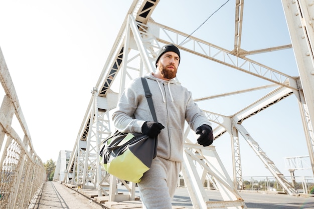 Portret van een serieuze bebaarde atleet met sporttas die langs de stadsbrug loopt