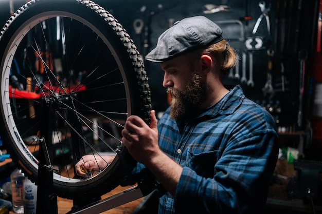 Portret van een serieuze bebaarde fietsende monteur man die fietswiel spaakt met fietsspaaksleutel in fietsenmaker met donker interieur Concept van professioneel onderhoud van fietstransport