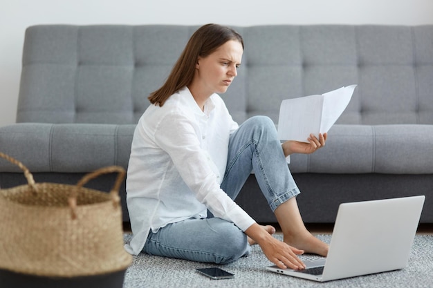 Portret van een serieuze, geconcentreerde vrouw met een wit overhemd en een spijkerbroek die op de vloer in de buurt van de bank zit met een laptop die papier vasthoudt met online les of een taak