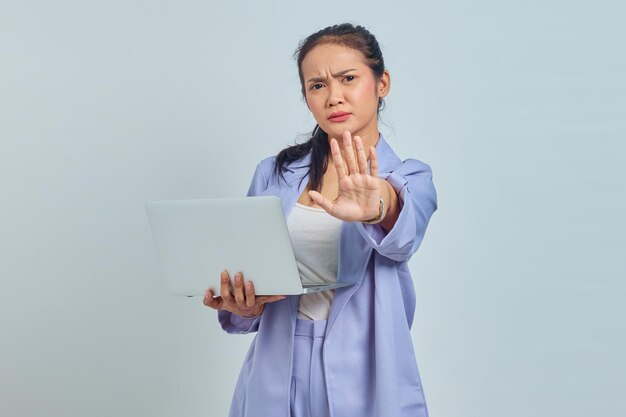 Portret van een serieuze jonge Aziatische vrouw die een laptop vasthoudt en met uitgestrekte hand staat met een stopgebaar dat op een witte achtergrond wordt geïsoleerd