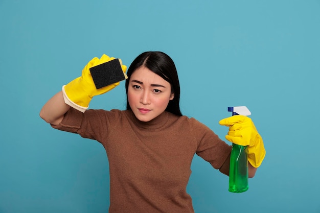 Portret van een serieuze jonge vrouw die gele handschoenen draagt met een spons wasmiddel sproeier geïsoleerd op een blauwe achtergrond, huisvrouw werknemer, schoonmaak huis concept, vrouwelijke boze gezichtsuitdrukking