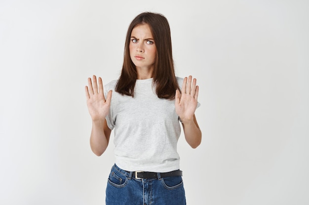 Portret van een serieuze jonge vrouw die met uitgestrekte hand staat en een stopgebaar toont dat over een witte muur wordt geïsoleerd, sterk weigert en nee zegt