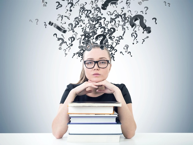 Foto portret van een serieuze jonge vrouw met een bril die vooruit kijkt en met haar kin op een stapel boeken zit. een grijze muurachtergrond met vraagtekens