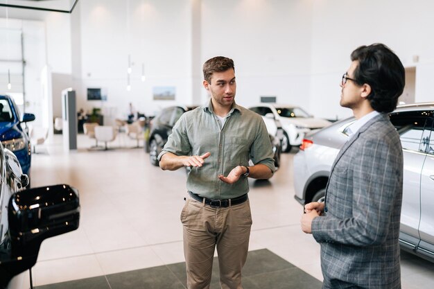 Portret van een serieuze mannelijke klant die met een professionele autodealer praat in een zakelijk pak in een autodealer die over auto's praat en naar een luxe nieuw model kijkt