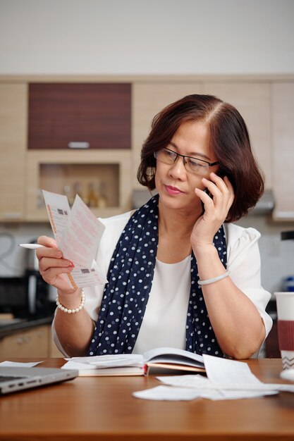 Portret van een serieuze oudere vrouw die aan de telefoon praat en rekeningen controleert terwijl ze aan de keukentafel zit