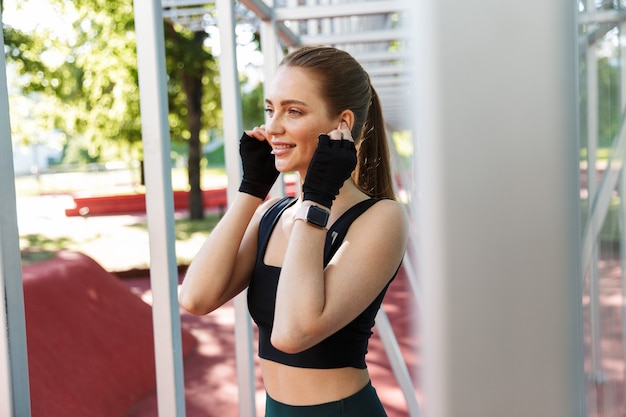 Portret van een slanke jonge vrouw in trainingspak die oordoppen draagt en training doet met een horizontale metalen staaf op sportterrein in groen park