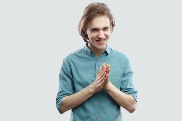 Portret van een sluwe knappe langharige blonde jongeman in een blauw casual shirt die staat en naar de camera kijkt met een bitchy plan. indoor studio opname, geïsoleerd op lichtgrijze achtergrond.