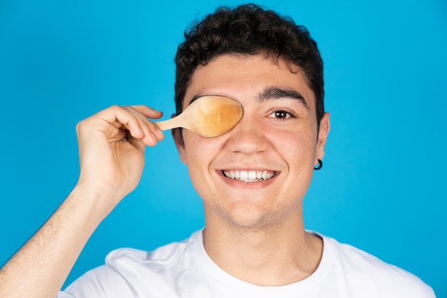 Portret van een Spaanse tienerjongen die een oog bedekt met een houten lepel die op een blauwe achtergrond wordt geïsoleerd