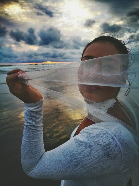 Portret van een speelse bruid met een sluier op het strand tegen bewolkte lucht tijdens de zonsondergang