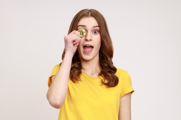 Portret van een speelse opgewonden vrouw van jonge leeftijd met een charmante glimlach die een geel T-shirt draagt dat één oog bedekt met gouden bitcoin, cryptocurrency. Indoor studio opname geïsoleerd op een grijze achtergrond.