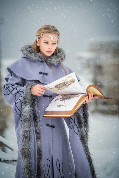 Portret van een sprookjesachtig elfmeisje met een magisch boek in haar handen tegen de achtergrond van de winternatuur en een magisch fort