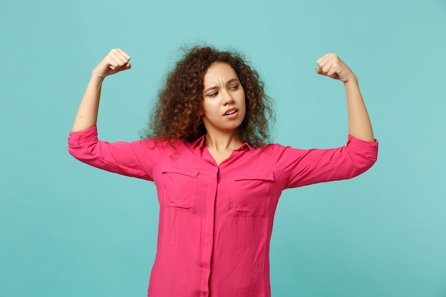 Portret van een sterk Afrikaans meisje in roze casual kleding met biceps, spieren geïsoleerd op een blauwe turquoise muurachtergrond in de studio. Mensen oprechte emoties, lifestyle concept. Bespotten kopie ruimte.