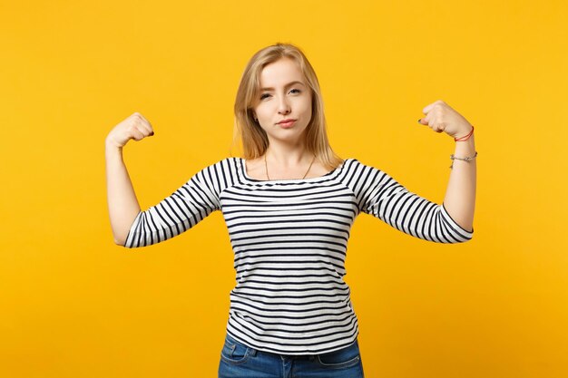 Portret van een sterke jonge vrouw in gestreepte kleding op zoek naar een camera met biceps, spieren geïsoleerd op een geeloranje achtergrond in de studio. Mensen oprechte emoties, lifestyle concept. Bespotten kopie ruimte.