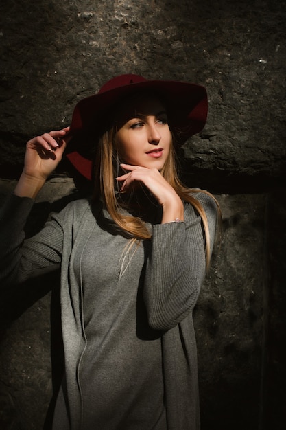 Foto portret van een stijlvol blond model met een grijze warme jurk en een rode hoed. vrouw poseren in zonlicht en schaduw op straat