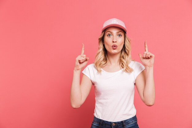 Portret van een stijlvolle blonde vrouw met een casual t-shirt en een pet die met de vingers naar boven wijst op copyspace geïsoleerd over roze muur