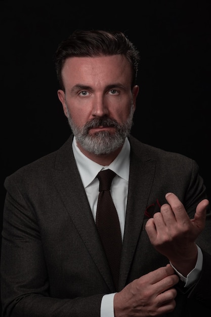 Portret van een stijlvolle elegante senior zakenman met een baard en casual zakelijke kleding in fotostudio geïsoleerd op een donkere achtergrond die het pak aanpast