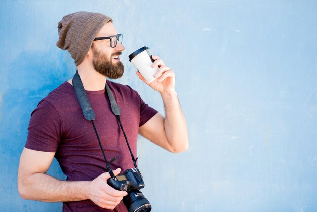 Portret van een stijlvolle fotograaf casual gekleed in een t-shirt en hoed met camera en koffie op de blauwe achtergrond