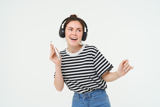 Portret van een stijlvolle jonge vrouw die in draadloze koptelefoon danst met behulp van koptelefoon en mobiele telefoon