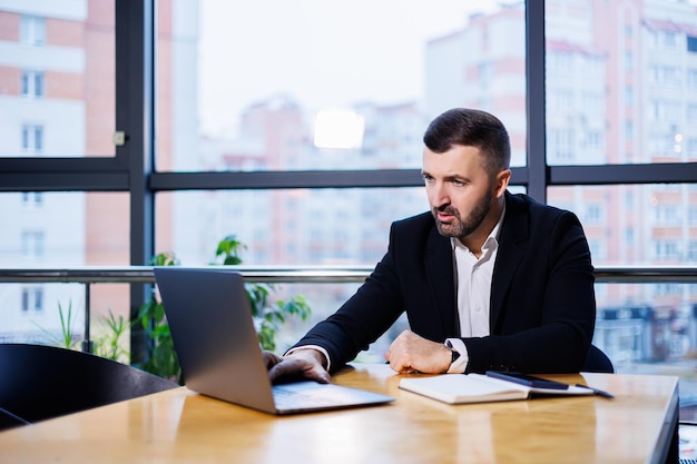 Portret van een stijlvolle jonge zakenman, in een groot modern kantoor hoog op een bovenverdieping, op zoek naar een laptop. Zakenman bij freelancer finance bureau