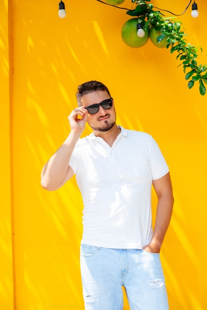 Portret van een stijlvolle jongeman met een baard in zonnebril en een wit t-shirt op straat tegen een felgele muur, lifestyle. Stedelijke stijl.