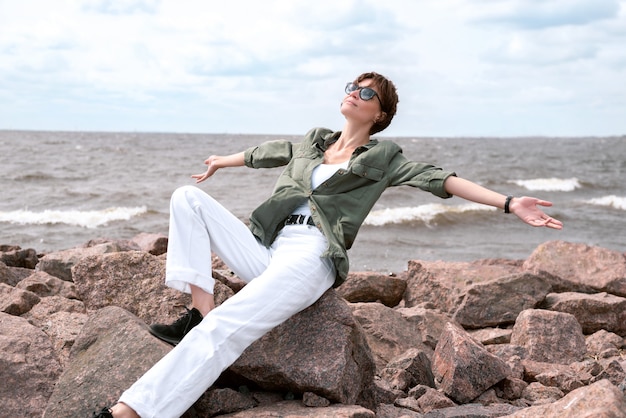 Portret van een stijlvolle vrouw zittend op stenen in de baai. Winderig weer. Geluk, vrijheid en zorgeloosheid. toeristisch concept